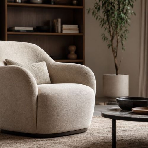 Poltrona di design comoda in tessuto bouclé beige, inserita in un soggiorno elegante con tavolino basso, libreria in legno, pianta e tende chiare.
